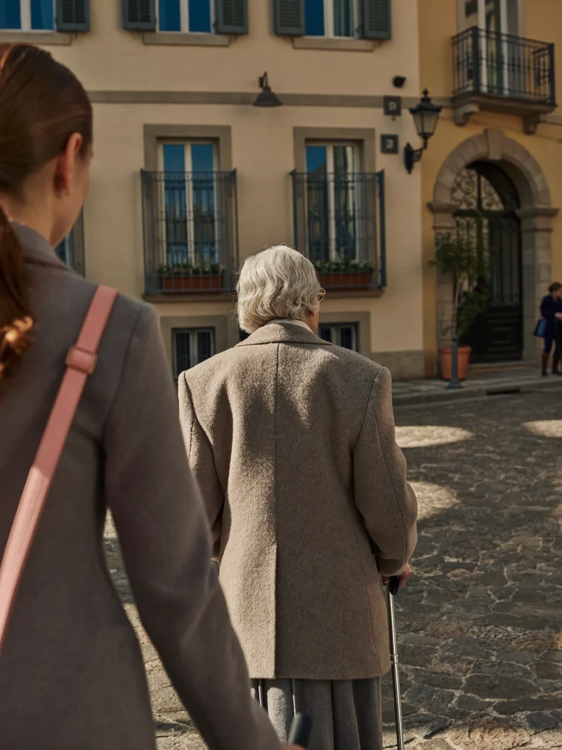 Un'accompagnatrice cammina accanto a una signora anziana verso l'ingresso di un ufficio locale.